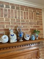 Front-facing shot of six Delft pottery pieces displayed on wooden mantel with brick wall background.