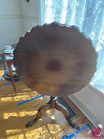 Front view of wooden pedestal table standing with scalloped round top face visible, showing wood grain and scratches.