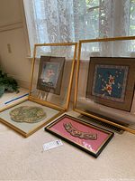 Four framed Asian fabric art pieces shown leaning against a wall near a window with lace curtains; includes two square blue floral pieces, oval patterned fabric, and an embroidered pink panel.