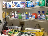 Wide view of laundry room shelves lined with various cleaning chemicals, detergents, 12-roll pack of Charmin toilet paper, and cleaning tools including brooms, mop, and sponges on washing machines below.