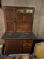 Front view of oak Hoosier style cabinet showing slag glass door inserts on upper doors, one large left vertical door, bread drawer roll-top in middle, and three drawers on right side with metal work surface.