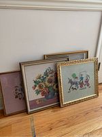 Four framed needlework pieces leaning against a wall on a hardwood floor, showing a variety of designs including floral and carriage scenes.
