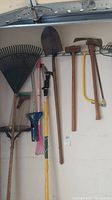 Photo showing various garden tools hanging on a wall including a large leaf rake, shovel, sledgehammer, hacksaw, broom, and telescoping pole pruner