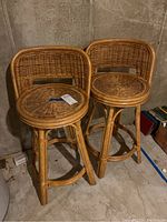 Two matching rattan swivel stools with woven seats and backs, standing on a basement concrete floor near a concrete wall.