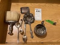 Seven sterling silver pieces arranged on wood surface: brush, tray, strainer spoon, two spoons, cup, thimble