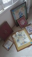 Overview of all six framed floral textile art pieces arranged on floor against wall, showing varied frame styles and floral embroidery designs.