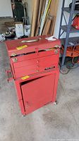 Front view of red metal tool chest with top compartment closed and bottom compartment door shut, showing wear and wheels.
