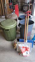 Photo showing two trash cans with lids (green and brown), two gray recycling bins, three snow shovels standing upright behind bins, bags of ice melt and water softener crystals on floor in garage setting.