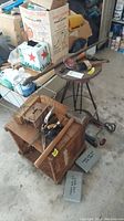 Wide shot showing wooden box, stool, assortment of tools including sledgehammer, ice tongs, hand plane, small hand tools, and first aid kits on concrete garage floor.