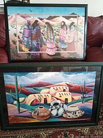 Photo showing both framed indigenous art prints on a red sofa with a glass coffee table in front - one print features five indigenous women in traditional dress with desert and mountain background, the other print shows adobe houses, cacti, and pottery.