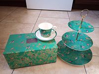 Tea cup, saucer, three-tier cake stand, and decorative box all displaying turquoise background with pink and dark blue birds and gold accents
