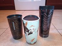 Three Starbucks drinkware items: two 6-inch mugs and one 8-inch tumbler arranged on tile floor