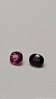 Two cushions cut natural sapphires, one bright pink and one deep purple, photographed side by side with light reflections visible on facets.