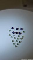 A collection of 19 loose gemstones arranged in rows on a flat surface, featuring deeper blue sapphires and lighter green alexandrites