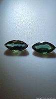 Two matched marquis cut sapphires in green blue tones, shown side by side on a plain surface under light to reveal faceting and color.