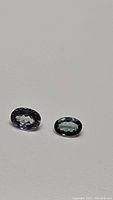 Two oval cut natural tanzanite gemstones on a light grey background, showing purple-blue color with green tone and good clarity.