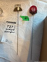 Two antique hat pins on a white textured fabric background, one with a filigree base and the other with a red carved component. Green sticker marked 10 inches length.