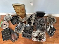 Overview of assortment of metal and cast iron bakeware pieces including trays, pans, muffin tins, racks, utensils, and cookie cutters arranged on floor.
