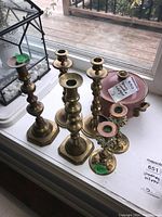 Photo showing six brass candle holders of varying designs and heights arranged on a windowsill next to a brass bowl