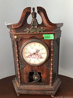 Front view of wooden quartz mantle clock showing Roman numeral clock face, decorative metal corners, finial, and pendulum window.