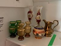 Photo showing six vases and two pitchers on a shelf: two green glass with gold accents, two white opaque glass with maroon and gold details, one small Wade ceramic pitcher with deer painting, one gold-tone metal pitcher.