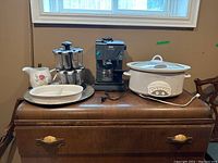 DeLonghi cappuccino maker, spice rack, ceramic pitcher, divided baking dish, aluminum tray, and crock pot displayed on wooden furniture under window.