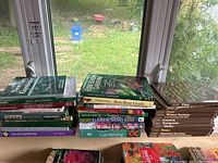 Photo showing stacks of gardening books on a window ledge with outdoor view in background. Titles about gardening topics visible including The New City Gardener, Small-Space Gardens, and various others.