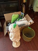 Photo showing the full lot including a decorative garden figure, a box containing various gardening supplies and a green ceramic planter.