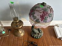 Photo of a metal and glass hurricane oil lamp with tall glass chimney next to a table lamp with a grapevine decorated glass shade and a resin base showing both lamps in full view