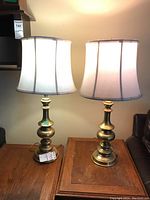 Two brass table lamps with white fabric lampshades standing on a wooden surface, lit from above.