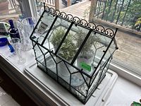 Front and side view of metal and glass miniature greenhouse containing faux plants and decorative white stones inside, placed on a windowsill near glassware.
