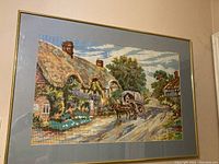 Framed needlepoint needlework showing a thatched roof cottage with flowering vines and a horse-drawn wagon on a country lane under a partly cloudy sky.