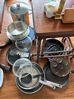 Overview of assorted stainless steel and cast iron cookware including pots, frying pans, glass lids, and cast iron muffin pan.