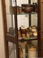 Three shelves in a glass cabinet filled with 19 pottery pieces of varying sizes and colors including brown, tan, and two prominent red vases.
