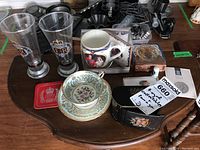 Overview photo of entire lot on a round wooden table showing all items together including glasses, cups, coin case, trinket box, and coaster