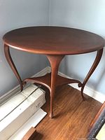 Side view of oval wooden side table showing three curved legs and lower triangular corner shelf, wood surface visible with slight scratches.
