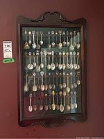 Front view of wood veneer display case holding multiple rows of collector spoons with varied decorative handles.