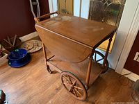 Side angle of wooden tea cart showing closed drop leaves, spoked wooden wheels, lower shelf, and handle.