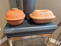 Two terra cotta baking dishes with lids placed on a stove top, showing shape and color