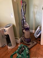Front-left side view of upright Dyson Ball 25 Animal vacuum cleaner with visible hose attachments and clear dust bin, standing next to a heater fan and iron board.