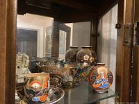 Display shelf with assorted vintage Japanese Satsuma pottery and decorative ceramics inside a glass cabinet, showing multiple hand-painted vases and bowls with traditional Japanese motifs.