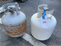 Two propane tanks side by side on pavement. Left tank is gray and rusty while right tank is white with a blue tag.