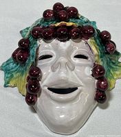 Front view of ceramic Bacchus mask showing facial features and grape cluster decoration on forehead and cheeks.