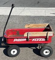 Side view of vintage red Radio Flyer wagon showing red metal body, wooden side rails, and large black rubber wheels with white hubs.