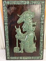 Full frontal photo showing wooden panel with greenish resin Aztec/Mayan figure and ornate border against dark wood background.