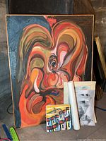 Large abstract painting on canvas with swirling warm colors framed in wood, with small cityscape painting and rolled artworks leaning against it.