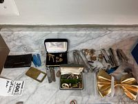 Overview of assorted men's accessories including tie clips, cuff links, a gold bow tie, metal nail clippers and a lighter on a marble surface.