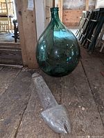 Photo showing large green-tinted glass Dame Jeanne vase with rounded shape and narrow neck, next to long metal and wood fish sculpture lying on wooden floor.
