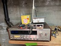 Wide view of vintage electronics on wood surface against concrete wall including Sylvania receiver, Sony alarm radio box, small silver device with antenna, and power adapter with cords.