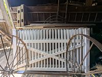 White painted wooden picket fencing panels with crisscross support detail leaning against a wall with wooden posts and planks behind them, in a garage, showing usage marks and dirt.